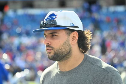 Ein Mann mit Bart und langen Haaren trägt ein graues T-Shirt mit einem Büffel-Logo, das an Matt Milanos Teamgeist erinnert. Er trägt eine weiß-blaue Kappe mit einer Sonnenbrille auf der Krempe, vor einer Stadionkulisse voller verschwommener Zuschauer bei einem spannenden Sportereignis. Diese Beschreibung wurde mit der FootballR KI automatisch generiert.