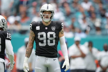 NFL Free Agency - Auf dem Spielfeld steht ein Footballspieler, Maxx Crosby, der als Teil der Las Vegas Raiders eine schwarz-silberne Uniform mit der Nummer 98 trägt. Er hat Tätowierungen auf seinem rechten Arm und trägt einen Helm mit Visier. Im Hintergrund sind unscharfe Zuschauer und Teammitglieder zu sehen. Diese Beschreibung wurde mit der FootballR KI automatisch generiert.