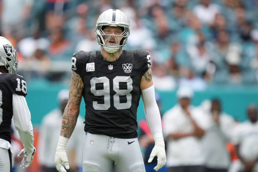 NFL Free Agency - Auf dem Spielfeld steht ein Footballspieler, Maxx Crosby, der als Teil der Las Vegas Raiders eine schwarz-silberne Uniform mit der Nummer 98 trägt. Er hat Tätowierungen auf seinem rechten Arm und trägt einen Helm mit Visier. Im Hintergrund sind unscharfe Zuschauer und Teammitglieder zu sehen. Diese Beschreibung wurde mit der FootballR KI automatisch generiert.