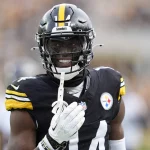 George Pickens im schwarz-gelben Trikot der Pittsburgh Steelers lächelt auf dem Spielfeld. Er trägt einen schwarzen Helm mit Visier und hält einen Mundschutz in der Hand, im Hintergrund sind unscharf jubelnde Fans zu sehen. Die Trikotnummer „14“ ist zu sehen. Diese Beschreibung wurde mit der FootballR KI automatisch generiert.