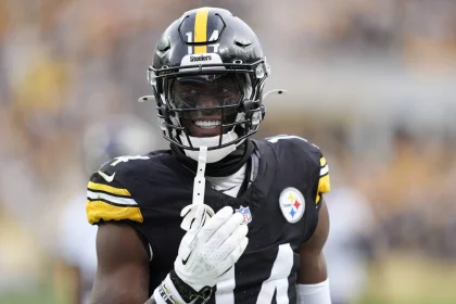 George Pickens im schwarz-gelben Trikot der Pittsburgh Steelers lächelt auf dem Spielfeld. Er trägt einen schwarzen Helm mit Visier und hält einen Mundschutz in der Hand, im Hintergrund sind unscharf jubelnde Fans zu sehen. Die Trikotnummer „14“ ist zu sehen. Diese Beschreibung wurde mit der FootballR KI automatisch generiert.