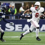 James Conner, ein Footballspieler in weißer Uniform, rennt während eines Spiels mit dem Ball, verfolgt von einem Spieler in Dunkelblau und Grün. Auf dem leuchtend grünen Feld mit Zuschauern in den Farben seines Teams wirkt Conner hochkonzentriert und hält den Ball fest, während er nach vorne stürmt. Diese Beschreibung wurde mit der FootballR KI automatisch generiert.