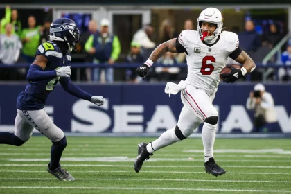 James Conner, ein Footballspieler in weißer Uniform, rennt während eines Spiels mit dem Ball, verfolgt von einem Spieler in Dunkelblau und Grün. Auf dem leuchtend grünen Feld mit Zuschauern in den Farben seines Teams wirkt Conner hochkonzentriert und hält den Ball fest, während er nach vorne stürmt. Diese Beschreibung wurde mit der FootballR KI automatisch generiert.