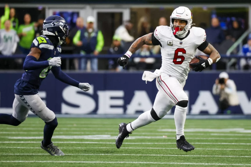 James Conner, ein Footballspieler in weißer Uniform, rennt während eines Spiels mit dem Ball, verfolgt von einem Spieler in Dunkelblau und Grün. Auf dem leuchtend grünen Feld mit Zuschauern in den Farben seines Teams wirkt Conner hochkonzentriert und hält den Ball fest, während er nach vorne stürmt. Diese Beschreibung wurde mit der FootballR KI automatisch generiert.