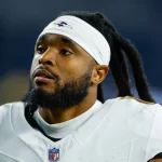 Auf dem Feld steht ein Footballspieler mit Bart und langen Dreadlocks, der an Diontae Johnson erinnert. Er trägt ein weißes Trikot mit einem lila-schwarzen Logo, ergänzt durch ein weißes Stirnband und Schulterpolster. Der Hintergrund ist unscharf und hebt den nachdenklichen Gesichtsausdruck des Spielers hervor. Diese Beschreibung wurde mit der FootballR KI automatisch generiert.