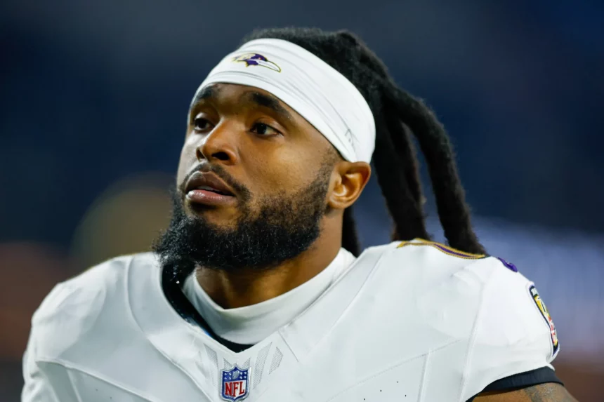 Auf dem Feld steht ein Footballspieler mit Bart und langen Dreadlocks, der an Diontae Johnson erinnert. Er trägt ein weißes Trikot mit einem lila-schwarzen Logo, ergänzt durch ein weißes Stirnband und Schulterpolster. Der Hintergrund ist unscharf und hebt den nachdenklichen Gesichtsausdruck des Spielers hervor. Diese Beschreibung wurde mit der FootballR KI automatisch generiert.