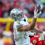 Brock Bowers - In einem spannenden NFL Recap Moment streckt sich ein Footballspieler in weißer Uniform und silbernem Helm, Nummer 89, für einen einhändigen Fang aus. Neben ihm ist ein gegnerischer Spieler in Rot teilweise zu sehen, während verschwommene Stadiontribünen voller rot gekleideter Zuschauer das Playoff-Szenario verfolgen. Diese Beschreibung wurde mit der FootballR KI automatisch generiert.