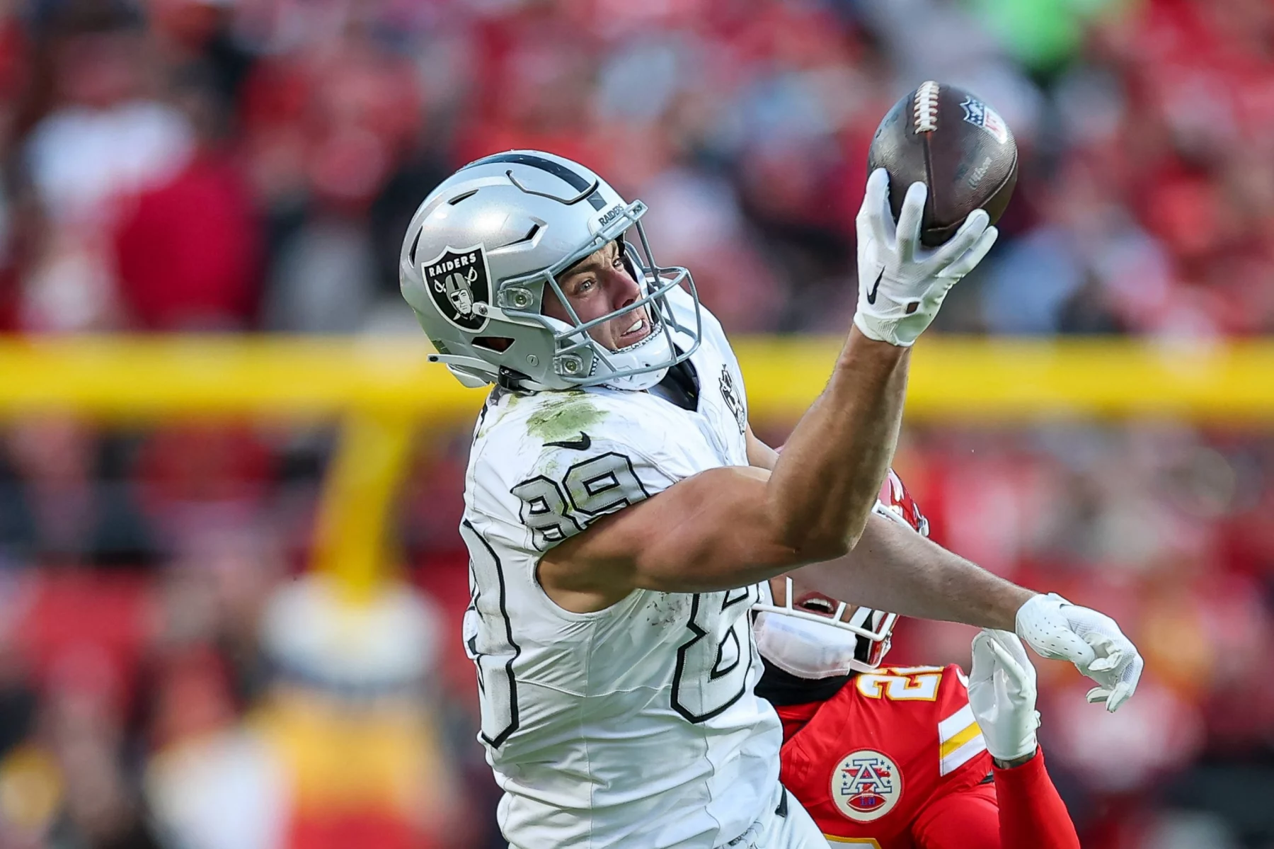 Brock Bowers - In einem spannenden NFL Recap Moment streckt sich ein Footballspieler in weißer Uniform und silbernem Helm, Nummer 89, für einen einhändigen Fang aus. Neben ihm ist ein gegnerischer Spieler in Rot teilweise zu sehen, während verschwommene Stadiontribünen voller rot gekleideter Zuschauer das Playoff-Szenario verfolgen. Diese Beschreibung wurde mit der FootballR KI automatisch generiert.