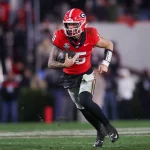 Carson Beck, der ein rotes Trikot mit der Nummer 5 trägt, sprintet mit einem schwarzen Football in der Hand über das Feld. Sein silberner Helm glänzt unter den Stadionlichtern, während er in schwarzen Hosen und einem weißen Ärmel läuft. Die Zuschauer und die anderen Spieler verschwimmen im Hintergrund und sind fasziniert von seiner Leistung. Diese Beschreibung wurde mit der FootballR KI automatisch generiert.