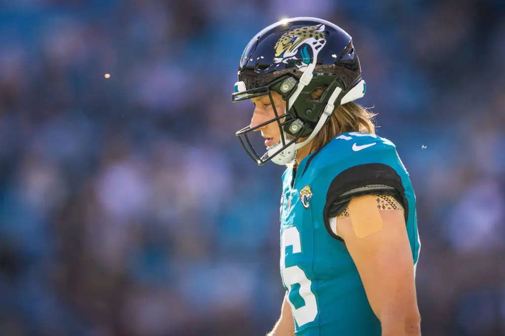Trevor Lawrence in einem blaugrĂŒnen Trikot und einem schwarzen Helm mit einem Jaguar-Logo steht auf dem Feld. Sein langes Haar flieĂt unter dem Helm hervor. An seinem rechten Arm befindet sich ein kleiner Verband. Im Hintergrund ist ein verschwommenes Stadion mit blauen Sitzen zu sehen, was auf eine Spielsituation schlieĂen lĂ€sst. Diese Beschreibung wurde mit der FootballR KI automatisch generiert.