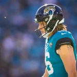 Trevor Lawrence in einem blaugrünen Trikot und einem schwarzen Helm mit einem Jaguar-Logo steht auf dem Feld. Sein langes Haar fließt unter dem Helm hervor. An seinem rechten Arm befindet sich ein kleiner Verband. Im Hintergrund ist ein verschwommenes Stadion mit blauen Sitzen zu sehen, was auf eine Spielsituation schließen lässt. Diese Beschreibung wurde mit der FootballR KI automatisch generiert.
