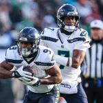 Zwei Spieler der Seattle Seahawks sind auf dem Feld in Aktion. Der Spieler links, Kenneth Walker III, hält den Ball fest und senkt seinen Körper zum Laufen, während der Spieler rechts bereit zu sein scheint, zu assistieren. Beide tragen weiße Trikots und dunkle Helme. Im Hintergrund ist ein Schiedsrichter zu sehen. Diese Beschreibung wurde mit der FootballR KI automatisch generiert.