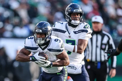 Zwei Spieler der Seattle Seahawks sind auf dem Feld in Aktion. Der Spieler links, Kenneth Walker III, hält den Ball fest und senkt seinen Körper zum Laufen, während der Spieler rechts bereit zu sein scheint, zu assistieren. Beide tragen weiße Trikots und dunkle Helme. Im Hintergrund ist ein Schiedsrichter zu sehen. Diese Beschreibung wurde mit der FootballR KI automatisch generiert.