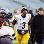 Ein Fußballspieler in weiß-gelber Uniform mit der Nummer 3 läuft mit breitem Lächeln über ein Feld. Er ist von Menschen umgeben, darunter ein Kameramann und ein Mann in dunklem Mantel und Mütze. Im Hintergrund ist ein leeres Stadion mit verschwommenen Zuschauern zu sehen. Diese Beschreibung wurde mit der FootballR KI automatisch generiert.