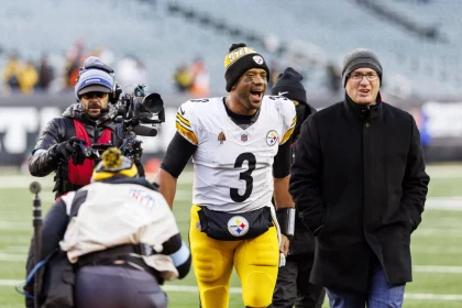 Ein Fußballspieler in weiß-gelber Uniform mit der Nummer 3 läuft mit breitem Lächeln über ein Feld. Er ist von Menschen umgeben, darunter ein Kameramann und ein Mann in dunklem Mantel und Mütze. Im Hintergrund ist ein leeres Stadion mit verschwommenen Zuschauern zu sehen. Diese Beschreibung wurde mit der FootballR KI automatisch generiert.