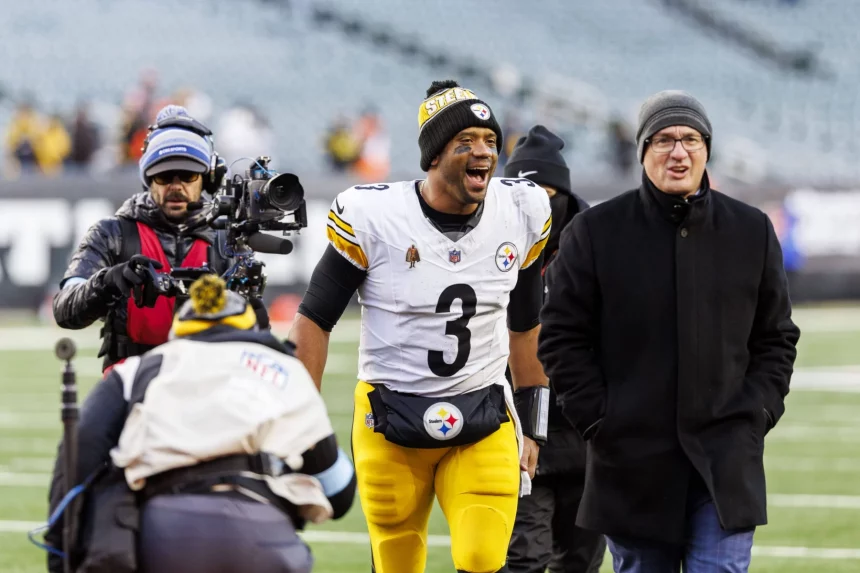 Ein Fußballspieler in weiß-gelber Uniform mit der Nummer 3 läuft mit breitem Lächeln über ein Feld. Er ist von Menschen umgeben, darunter ein Kameramann und ein Mann in dunklem Mantel und Mütze. Im Hintergrund ist ein leeres Stadion mit verschwommenen Zuschauern zu sehen. Diese Beschreibung wurde mit der FootballR KI automatisch generiert.