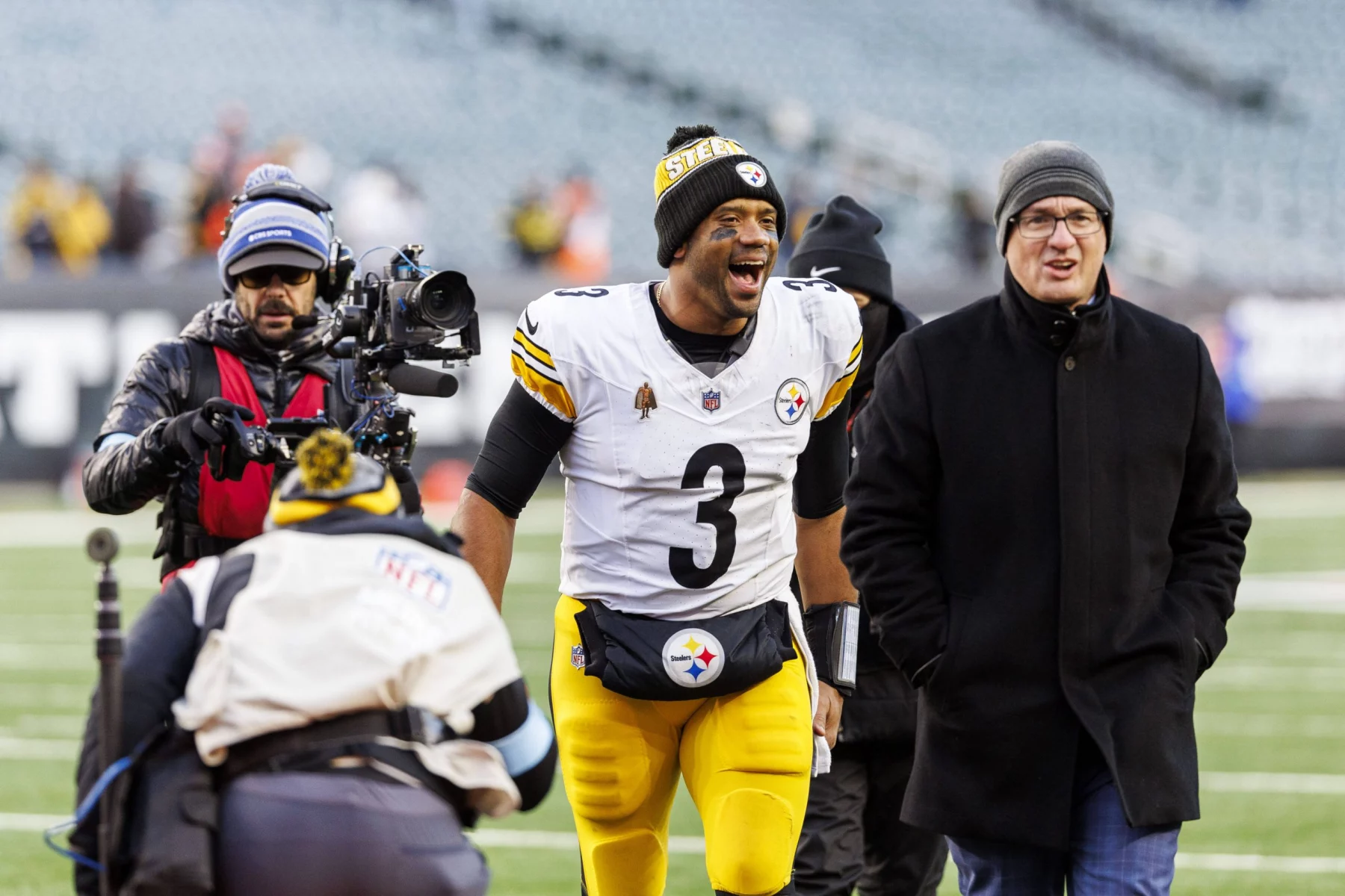 Ein Fußballspieler in weiß-gelber Uniform mit der Nummer 3 läuft mit breitem Lächeln über ein Feld. Er ist von Menschen umgeben, darunter ein Kameramann und ein Mann in dunklem Mantel und Mütze. Im Hintergrund ist ein leeres Stadion mit verschwommenen Zuschauern zu sehen. Diese Beschreibung wurde mit der FootballR KI automatisch generiert.