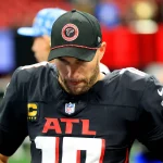 Ein Footballspieler, der in seiner Konzentration an Kirk Cousins erinnert, trägt auf dem Spielfeld ein schwarzes Trikot und eine schwarze Kappe der Atlanta Falcons. Das Trikot trägt die Nummer 10, während die Kappe das Logo der Falcons trägt. Mit den Händen in den Hüften und konzentrierter Haltung steht er vor einer verschwommenen Stadionkulisse. Diese Beschreibung wurde mit der FootballR KI automatisch generiert.