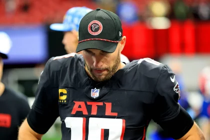 Ein Footballspieler, der in seiner Konzentration an Kirk Cousins erinnert, trägt auf dem Spielfeld ein schwarzes Trikot und eine schwarze Kappe der Atlanta Falcons. Das Trikot trägt die Nummer 10, während die Kappe das Logo der Falcons trägt. Mit den Händen in den Hüften und konzentrierter Haltung steht er vor einer verschwommenen Stadionkulisse. Diese Beschreibung wurde mit der FootballR KI automatisch generiert.