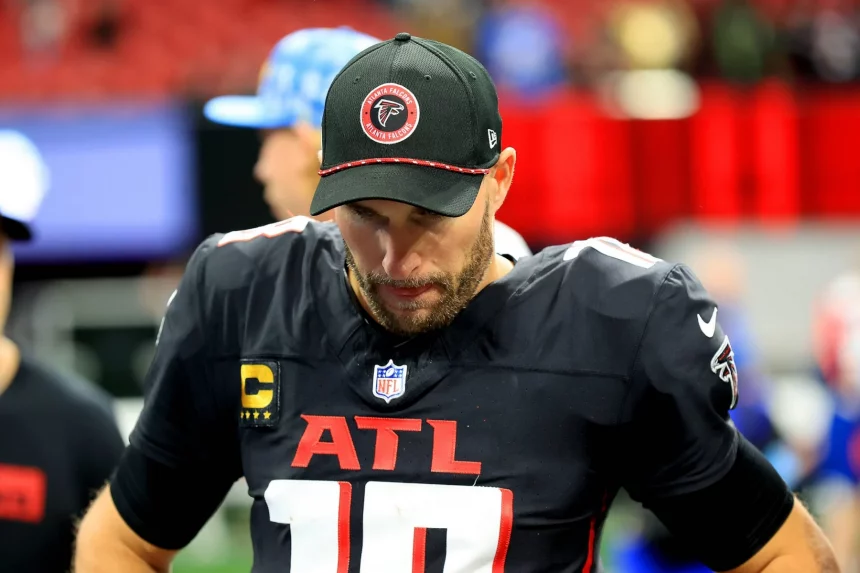 Ein Footballspieler, der in seiner Konzentration an Kirk Cousins erinnert, trägt auf dem Spielfeld ein schwarzes Trikot und eine schwarze Kappe der Atlanta Falcons. Das Trikot trägt die Nummer 10, während die Kappe das Logo der Falcons trägt. Mit den Händen in den Hüften und konzentrierter Haltung steht er vor einer verschwommenen Stadionkulisse. Diese Beschreibung wurde mit der FootballR KI automatisch generiert.