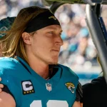 Ein Footballspieler mit langen Haaren, unverkennbar Trevor Lawrence, trägt ein blaugrünes Jacksonville Jaguars-Trikot mit der Nummer 16 und dem Kapitänsabzeichen und sitzt konzentriert auf einem Fahrzeug. Eine Person hinter ihm legt eine Hand auf seine Schulter. Im Hintergrund ist eine verschwommene Menschenmenge in einem Stadion zu sehen. Diese Beschreibung wurde mit der FootballR KI automatisch generiert.