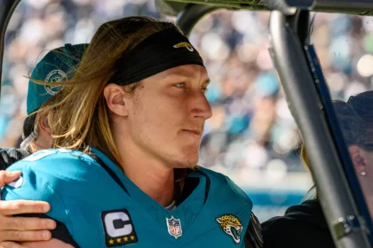Ein Footballspieler mit langen Haaren, unverkennbar Trevor Lawrence, trägt ein blaugrünes Jacksonville Jaguars-Trikot mit der Nummer 16 und dem Kapitänsabzeichen und sitzt konzentriert auf einem Fahrzeug. Eine Person hinter ihm legt eine Hand auf seine Schulter. Im Hintergrund ist eine verschwommene Menschenmenge in einem Stadion zu sehen. Diese Beschreibung wurde mit der FootballR KI automatisch generiert.