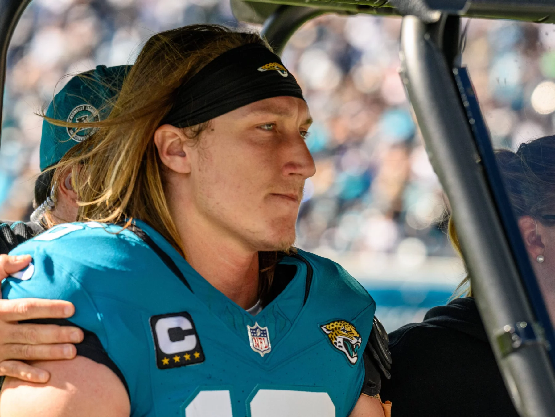Ein Footballspieler mit langen Haaren, unverkennbar Trevor Lawrence, trägt ein blaugrünes Jacksonville Jaguars-Trikot mit der Nummer 16 und dem Kapitänsabzeichen und sitzt konzentriert auf einem Fahrzeug. Eine Person hinter ihm legt eine Hand auf seine Schulter. Im Hintergrund ist eine verschwommene Menschenmenge in einem Stadion zu sehen. Diese Beschreibung wurde mit der FootballR KI automatisch generiert.