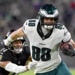 Ein Footballspieler, Dallas Goedert, trägt ein weiß-grünes Eagles-Trikot mit der Nummer 88 und rennt mit dem Ball unter seinem rechten Arm über das Feld. Ein Spieler der gegnerischen Mannschaft in schwarzer Uniform greift ihn von hinten an, um ihn vor dem Hintergrund einer verschwommenen Stadionkulisse zu tackeln. Diese Beschreibung wurde mit der FootballR KI automatisch generiert.