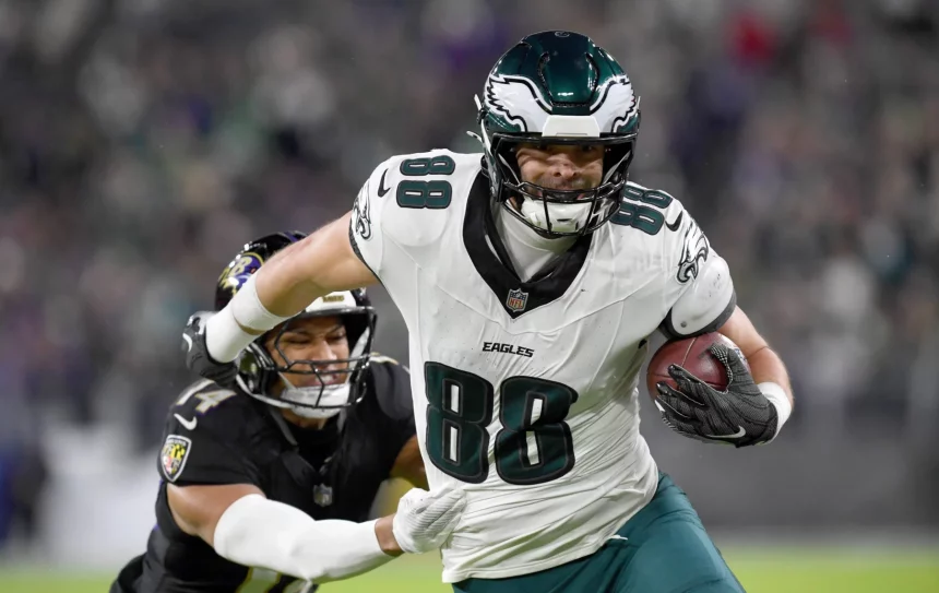 Ein Footballspieler, Dallas Goedert, trägt ein weiß-grünes Eagles-Trikot mit der Nummer 88 und rennt mit dem Ball unter seinem rechten Arm über das Feld. Ein Spieler der gegnerischen Mannschaft in schwarzer Uniform greift ihn von hinten an, um ihn vor dem Hintergrund einer verschwommenen Stadionkulisse zu tackeln. Diese Beschreibung wurde mit der FootballR KI automatisch generiert.