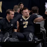 Taysom Hill in schwarz-goldener Uniform und mit Kapitänsabzeichen sitzt auf einem Krankenwagen. Er hebt eine behandschuhte Hand und grüßt die Menge. Ihre Mienen sind ruhig, im verschwommenen Hintergrund sind ein paar Personen zu sehen. Diese Beschreibung wurde mit der FootballR KI automatisch generiert.