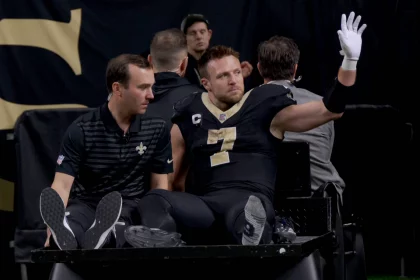 Taysom Hill in schwarz-goldener Uniform und mit Kapitänsabzeichen sitzt auf einem Krankenwagen. Er hebt eine behandschuhte Hand und grüßt die Menge. Ihre Mienen sind ruhig, im verschwommenen Hintergrund sind ein paar Personen zu sehen. Diese Beschreibung wurde mit der FootballR KI automatisch generiert.