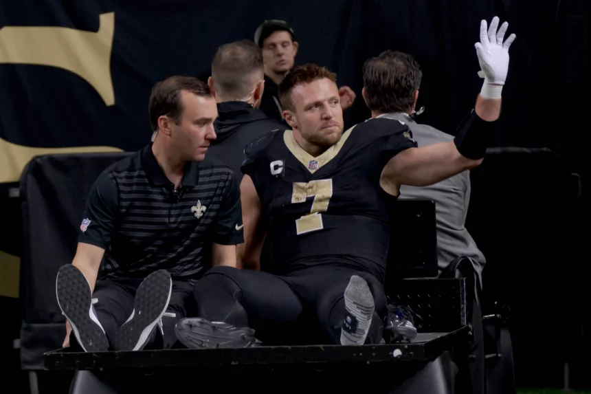 Taysom Hill in schwarz-goldener Uniform und mit Kapitänsabzeichen sitzt auf einem Krankenwagen. Er hebt eine behandschuhte Hand und grüßt die Menge. Ihre Mienen sind ruhig, im verschwommenen Hintergrund sind ein paar Personen zu sehen. Diese Beschreibung wurde mit der FootballR KI automatisch generiert.