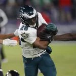 Saquon Barkley in einer weiß-grünen Uniform, der den Ball trägt, wird auf einem NFL-Feld von zwei Gegnern in schwarz-lila Uniformen angegriffen. Der Helm des Spielers trägt ein Flügelemblem. Die Action ist intensiv und konzentriert sich auf den entschlossenen Gesichtsausdruck des Spielers, während er sich durch das Tackling kämpft. Diese Beschreibung wurde mit der FootballR KI automatisch generiert.