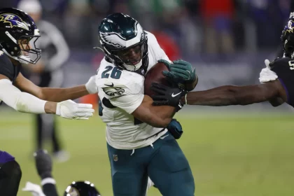 Saquon Barkley in einer weiß-grünen Uniform, der den Ball trägt, wird auf einem NFL-Feld von zwei Gegnern in schwarz-lila Uniformen angegriffen. Der Helm des Spielers trägt ein Flügelemblem. Die Action ist intensiv und konzentriert sich auf den entschlossenen Gesichtsausdruck des Spielers, während er sich durch das Tackling kämpft. Diese Beschreibung wurde mit der FootballR KI automatisch generiert.