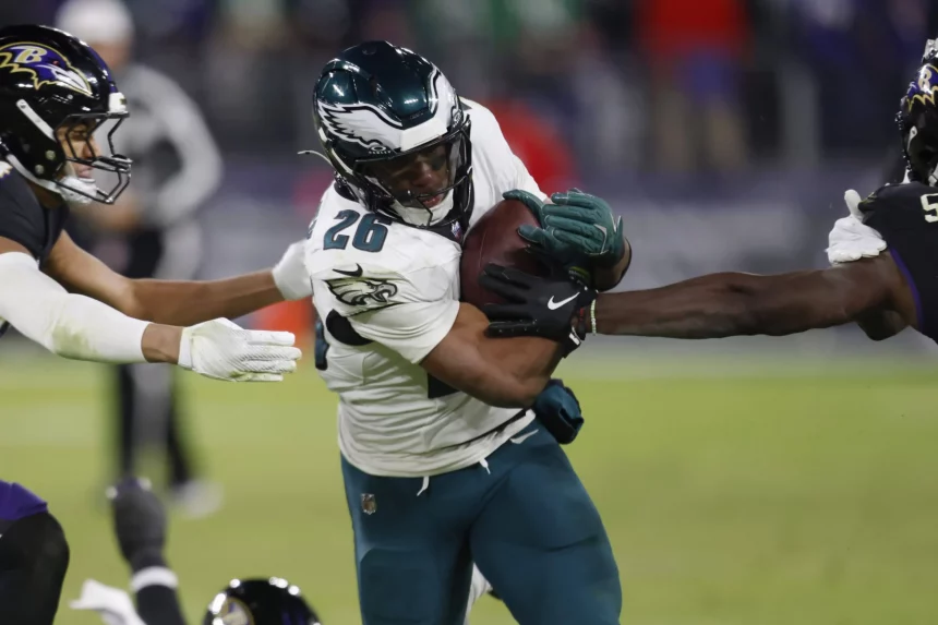 Saquon Barkley in einer weiß-grünen Uniform, der den Ball trägt, wird auf einem NFL-Feld von zwei Gegnern in schwarz-lila Uniformen angegriffen. Der Helm des Spielers trägt ein Flügelemblem. Die Action ist intensiv und konzentriert sich auf den entschlossenen Gesichtsausdruck des Spielers, während er sich durch das Tackling kämpft. Diese Beschreibung wurde mit der FootballR KI automatisch generiert.