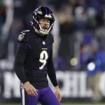 Ein Footballspieler in einem schwarzen Trikot mit der Nummer 9 und einem Ravens-Helm steht mit offenem Mund auf dem Feld. Im Hintergrund sind Fans und Stadionlichter zu sehen, die die intensive Spielatmosphäre der NFL einfangen, während Justin Tucker versucht, seine Formkrise zu überwinden. Diese Beschreibung wurde mit der FootballR KI automatisch generiert.