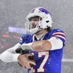 Ein Footballspieler in einem blauen Trikot mit der Nummer 17 steht im dichten Schnee auf einem Feld und hält einen Football in der Hand. Während das NFL Playoff Picture intensiver wird, trägt er einen weißen Helm mit klarem Visier vor verschwommenem Stadionlicht, während dichte Schneeflocken um ihn herum fallen. Diese Beschreibung wurde mit der FootballR KI automatisch generiert.