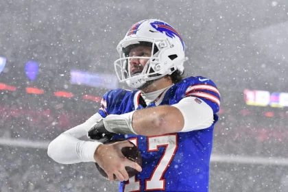 Ein Footballspieler in einem blauen Trikot mit der Nummer 17 steht im dichten Schnee auf einem Feld und hält einen Football in der Hand. Während das NFL Playoff Picture intensiver wird, trägt er einen weißen Helm mit klarem Visier vor verschwommenem Stadionlicht, während dichte Schneeflocken um ihn herum fallen. Diese Beschreibung wurde mit der FootballR KI automatisch generiert.