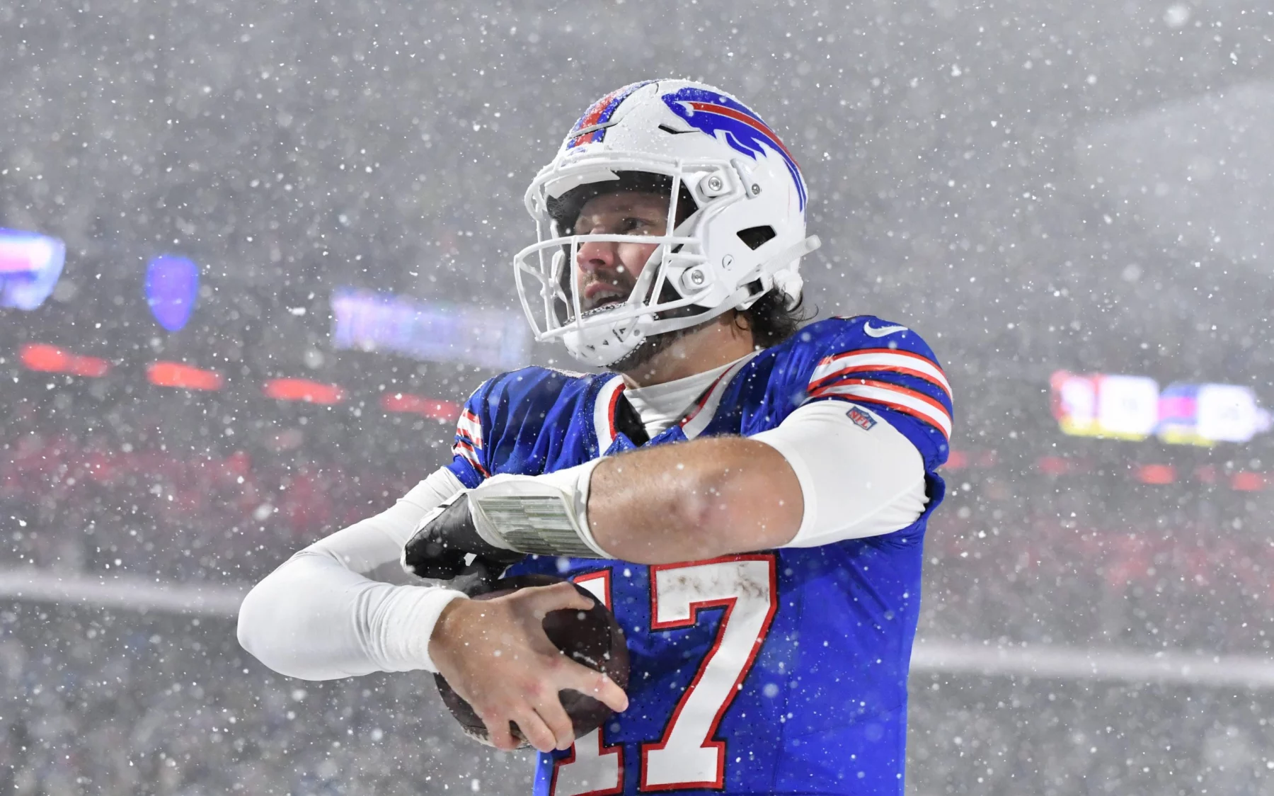 Ein Footballspieler in einem blauen Trikot mit der Nummer 17 steht im dichten Schnee auf einem Feld und hält einen Football in der Hand. Während das NFL Playoff Picture intensiver wird, trägt er einen weißen Helm mit klarem Visier vor verschwommenem Stadionlicht, während dichte Schneeflocken um ihn herum fallen. Diese Beschreibung wurde mit der FootballR KI automatisch generiert.