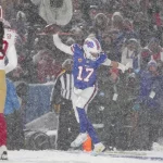 Ein Footballspieler in einer blauen Uniform, die an die Bills erinnert, springt in einem verschneiten Stadion, um seinen Sieg in Woche 14 der NFL zu feiern. Er hält einen Football in der Hand, während ein anderer Spieler in Rot und Gold in der Nähe zusieht. Es schneit heftig und schafft eine winterliche Atmosphäre. Im Hintergrund sind Zuschauer zu sehen. Diese Beschreibung wurde mit der FootballR KI automatisch generiert.
