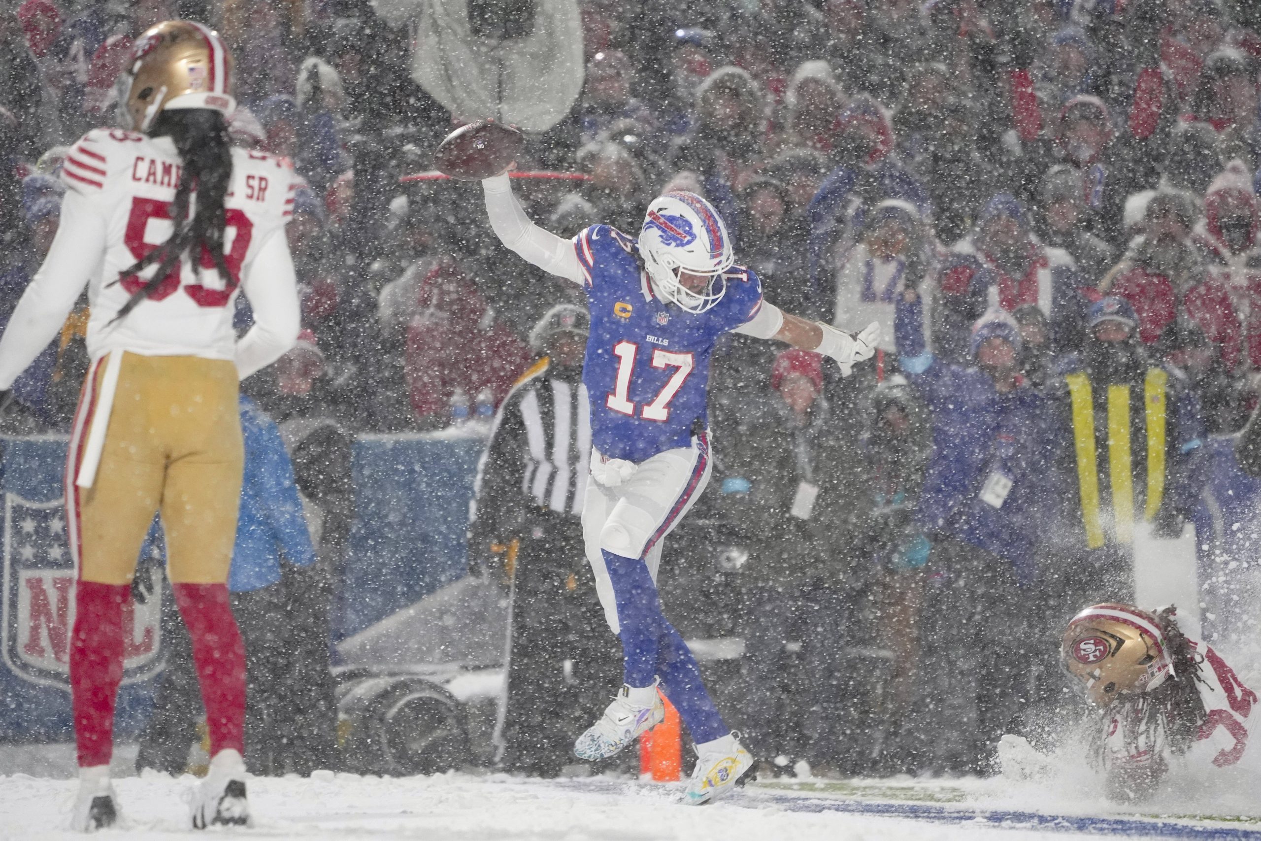 Ein Footballspieler in einer blauen Uniform, die an die Bills erinnert, springt in einem verschneiten Stadion, um seinen Sieg in Woche 14 der NFL zu feiern. Er hält einen Football in der Hand, während ein anderer Spieler in Rot und Gold in der Nähe zusieht. Es schneit heftig und schafft eine winterliche Atmosphäre. Im Hintergrund sind Zuschauer zu sehen. Diese Beschreibung wurde mit der FootballR KI automatisch generiert.