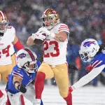 Christian McCaffrey, in einer weiß-roten Uniform, läuft mit dem Ball und weicht dabei gegnerischen Spielern in Blau und Rot aus. Schnee fällt auf das Feld und intensiviert die Atmosphäre. Als ein gegnerischer Spieler versucht, ihn zu tackeln, bleibt die Action dynamisch und intensiv. Diese Beschreibung wurde mit der FootballR KI automatisch generiert.