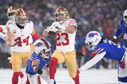 Christian McCaffrey, in einer weiß-roten Uniform, läuft mit dem Ball und weicht dabei gegnerischen Spielern in Blau und Rot aus. Schnee fällt auf das Feld und intensiviert die Atmosphäre. Als ein gegnerischer Spieler versucht, ihn zu tackeln, bleibt die Action dynamisch und intensiv. Diese Beschreibung wurde mit der FootballR KI automatisch generiert.