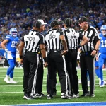 Eine Gruppe von Football-Schiedsrichtern versammelt sich auf dem Spielfeld. Sie tragen schwarz-weiß gestreifte Uniformen und schwarze Hüte und besprechen einen Spielzug. Mithilfe von Replay Assist sorgen sie für Genauigkeit. Sie stehen auf grünem Rasen, umgeben von Spielern der Detroit Lions und der Green Bay Packers, während das Stadionpublikum gespannt zusieht. Diese Beschreibung wurde mit der FootballR KI automatisch generiert.