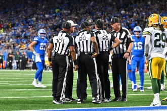 Eine Gruppe von Football-Schiedsrichtern versammelt sich auf dem Spielfeld. Sie tragen schwarz-weiß gestreifte Uniformen und schwarze Hüte und besprechen einen Spielzug. Mithilfe von Replay Assist sorgen sie für Genauigkeit. Sie stehen auf grünem Rasen, umgeben von Spielern der Detroit Lions und der Green Bay Packers, während das Stadionpublikum gespannt zusieht. Diese Beschreibung wurde mit der FootballR KI automatisch generiert.