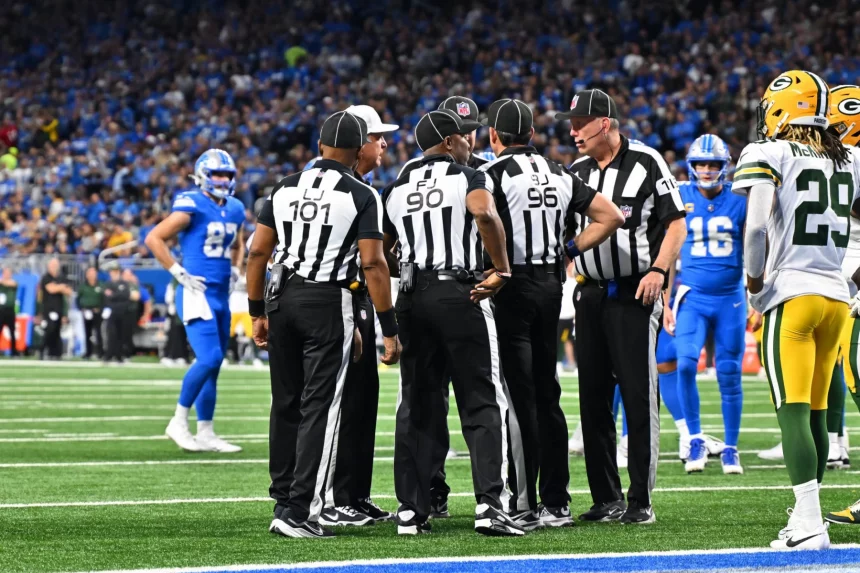 Eine Gruppe von Football-Schiedsrichtern versammelt sich auf dem Spielfeld. Sie tragen schwarz-weiß gestreifte Uniformen und schwarze Hüte und besprechen einen Spielzug. Mithilfe von Replay Assist sorgen sie für Genauigkeit. Sie stehen auf grünem Rasen, umgeben von Spielern der Detroit Lions und der Green Bay Packers, während das Stadionpublikum gespannt zusieht. Diese Beschreibung wurde mit der FootballR KI automatisch generiert.