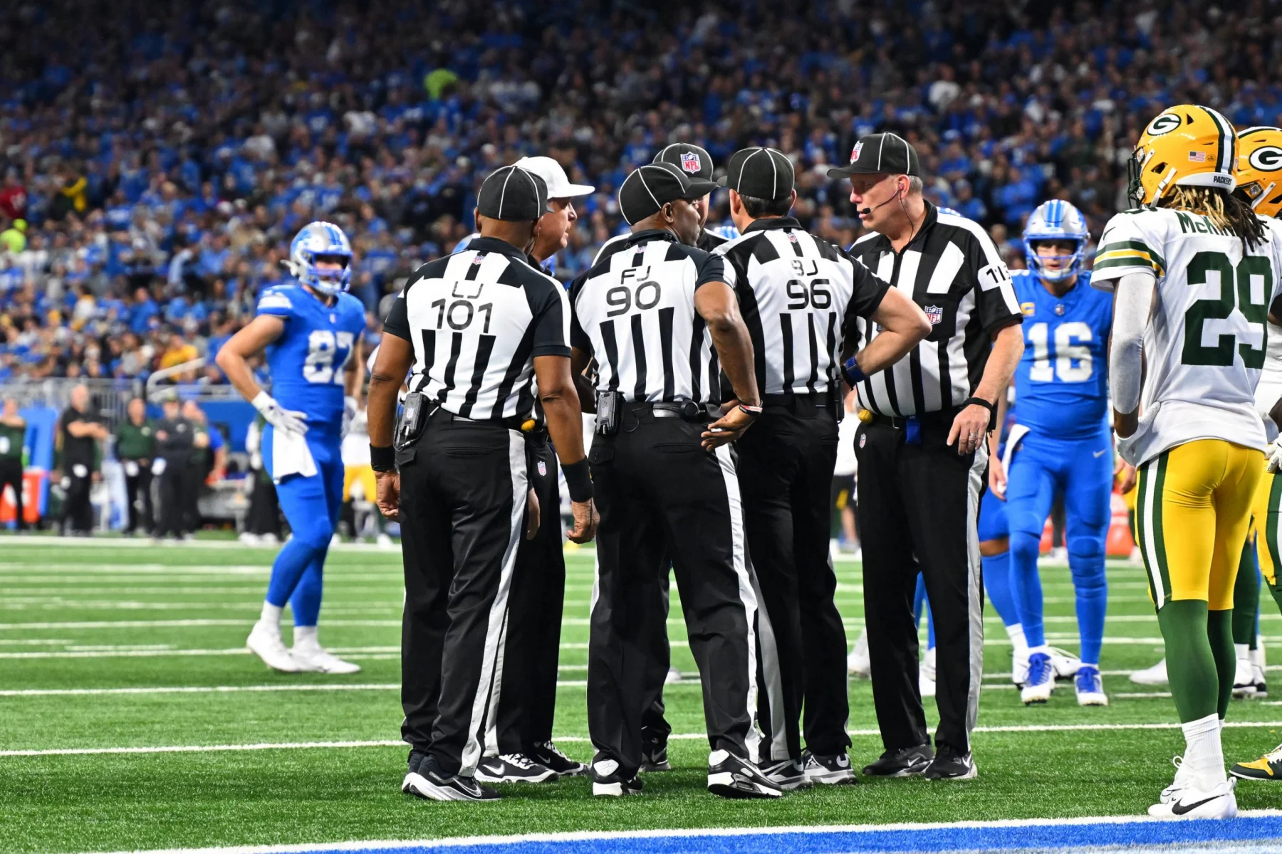 Eine Gruppe von Football-Schiedsrichtern versammelt sich auf dem Spielfeld. Sie tragen schwarz-weiß gestreifte Uniformen und schwarze Hüte und besprechen einen Spielzug. Mithilfe von Replay Assist sorgen sie für Genauigkeit. Sie stehen auf grünem Rasen, umgeben von Spielern der Detroit Lions und der Green Bay Packers, während das Stadionpublikum gespannt zusieht. Diese Beschreibung wurde mit der FootballR KI automatisch generiert.