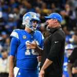 Ein Footballspieler in blauer Detroit-Lions-Uniform und Helm steht neben einem Trainer, der ein schwarzes Shirt und eine blaue Mütze trägt. Sie scheinen am Spielfeldrand eine Strategie zu besprechen. Im Hintergrund ist eine verschwommene Menschenmenge in einem Stadion zu sehen. Diese Beschreibung wurde mit der FootballR KI automatisch generiert.