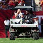 Ein Footballspieler, Raiders-Quarterback Aidan O'Connell, sitzt in einer weiß-schwarzen Uniform auf der Rückseite eines Wagens und bedeckt sein Gesicht mit den Händen, wahrscheinlich aufgrund einer Knieverletzung. Neben ihm steht eine weitere Person in Schwarz. Sie befinden sich auf einem Footballfeld mit einer Menschenmenge im Hintergrund, von denen einige rot gekleidet sind. Diese Beschreibung wurde mit der FootballR KI automatisch generiert.