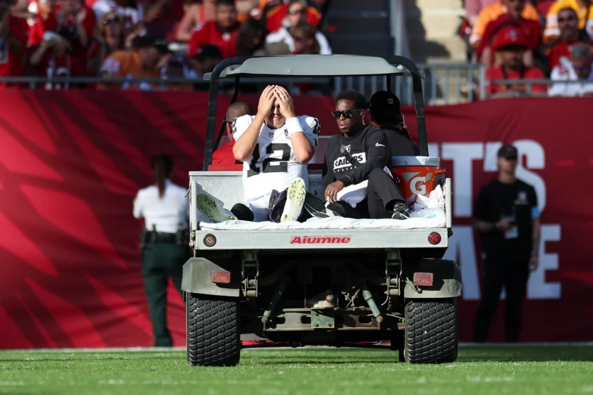 Ein Footballspieler, Raiders-Quarterback Aidan O'Connell, sitzt in einer weiß-schwarzen Uniform auf der Rückseite eines Wagens und bedeckt sein Gesicht mit den Händen, wahrscheinlich aufgrund einer Knieverletzung. Neben ihm steht eine weitere Person in Schwarz. Sie befinden sich auf einem Footballfeld mit einer Menschenmenge im Hintergrund, von denen einige rot gekleidet sind. Diese Beschreibung wurde mit der FootballR KI automatisch generiert.